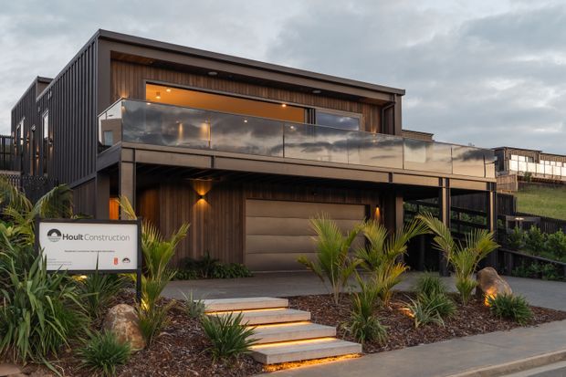 Exterior architectural photograph of a contemporary multi-level home with dark vertical cladding, warm timber-look wall panels, a broad cantilevered upper balcony with clear glass balustrade, recessed lighting, and a large garage door. A landscaped front garden with palms and illuminated steps leads to the entry, and a construction sign is visible in the foreground.