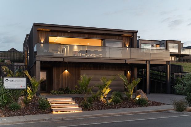 Exterior view of a contemporary multi-level residence at dusk, featuring dark timber-look cladding, a broad upper-floor balcony with glass balustrade, warm recessed lighting, large glazed openings, and landscaped planting along an illuminated front path. A covered lower-level area and driveway are visible.