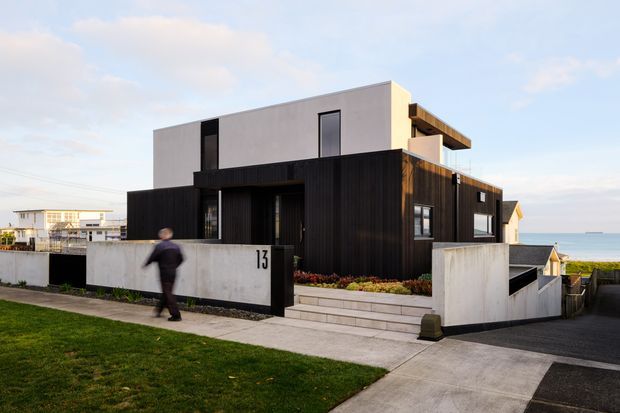 Street-facing exterior view of a contemporary multi-level coastal home. The building combines a dark vertically clad lower volume with a smooth light upper box, large dark-framed windows, a stepped entry path, low rendered boundary walls, and an integrated garage or driveway. The ocean is visible beyond the property.