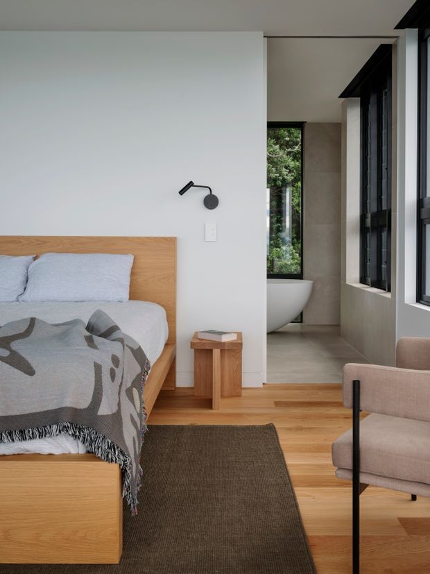 A calm contemporary bedroom with a light timber platform bed and headboard, pale bedding, a patterned throw, and a small timber bedside table. A partially open ensuite is visible beyond the dividing wall, containing a freestanding white bath, large neutral wall tiles, and dark-framed windows. Warm timber flooring, a dark textured rug, a black wall-mounted reading lamp, and a neutral upholstered armchair complete the room.