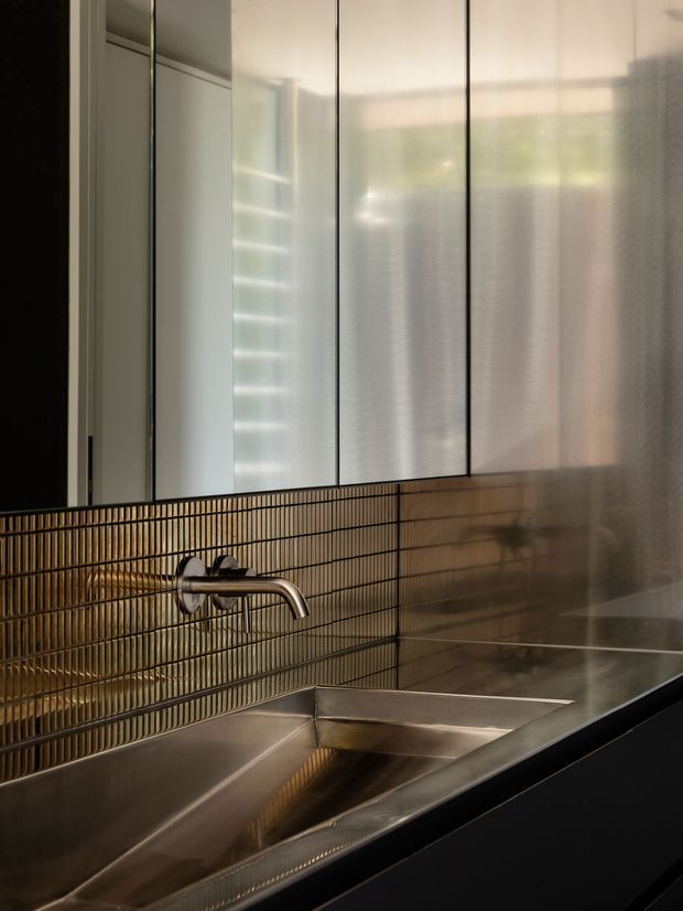 Close-up of a refined contemporary bathroom vanity featuring a large stainless-steel sink and minimalist wall-mounted brushed-metal tap. The backsplash is clad in narrow vertical bronze-brown tiles, with a full-width mirrored cabinet or mirror above reflecting a softly lit window and blinds. The composition emphasizes metallic surfaces, linear detailing, and warm ambient light.