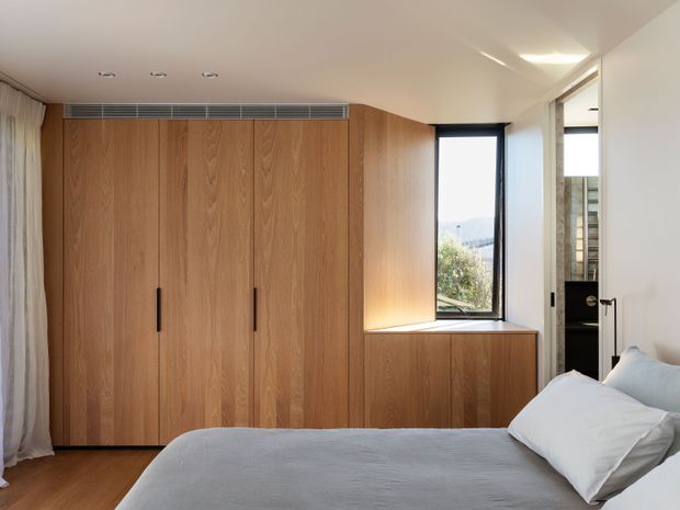 Contemporary bedroom with a large wall of warm timber built-in wardrobes and integrated low cabinetry beneath a narrow black-framed window. A partially visible ensuite is accessed through the open doorway at right. The room has a pale neutral ceiling and walls, timber flooring, soft white curtains, and a simple upholstered bed, creating a clean, warm, minimalist atmosphere.