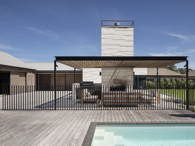 A contemporary single-storey residential exterior with a flat-roofed covered outdoor entertaining area beside a swimming pool. The home features a tall white textured masonry chimney, dark metal posts and railings, timber decking, timber-clad walls, and a slatted pergola roof casting patterned shadows. A lawn and neighboring structures are visible beyond the fenced terrace.