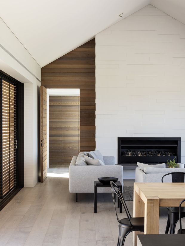 Bright contemporary living room with an open connection to a dining area. The room features a high sloping ceiling, pale timber flooring, a large white textured feature wall with a built-in black fireplace, warm horizontal timber wall panelling around a doorway, and expansive windows with dark timber blinds. Light grey sofas and black metal furniture create a clean, refined interior.