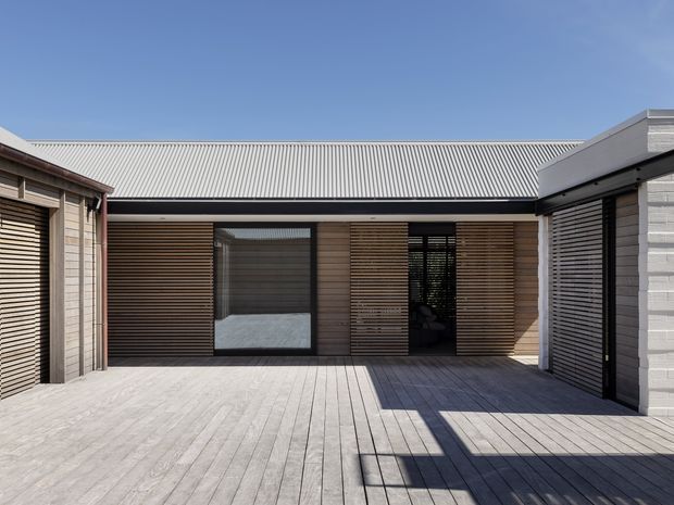 A modern single-storey courtyard-style building viewed from an expansive timber deck. The facade combines horizontal timber slats, dark-framed glazing and pale masonry walls, with a low-pitched corrugated metal roof and strong geometric shadows. The courtyard is enclosed by multiple connected wings and appears bright, private and minimalist.