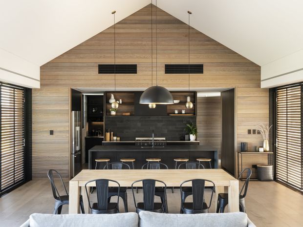 A warm, open-plan kitchen and dining area with a dramatic vaulted timber-lined gable wall and ceiling. The kitchen features dark fixed cabinetry, a dark tiled splashback, a long island with seating, integrated appliances and pendant lighting. In the foreground is a light timber dining table with black metal chairs, while timber venetian blinds frame the side windows and a sofa edge is visible at the bottom of the image.