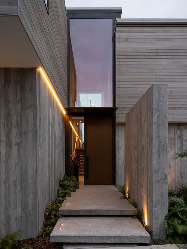 A dramatic modern residential entryway viewed from outside, approached by floating concrete steps and a narrow concrete path. The facade combines vertical timber cladding, textured concrete walls, dark metal-framed glazing, and a tall glazed volume above the entrance. Warm linear and low-level lighting highlights the walls and planted edges, creating a refined, moody arrival sequence.