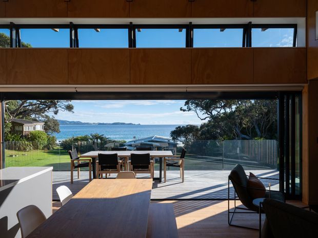 A bright, mid-century-inspired open-plan dining and living area opening onto a timber deck with frameless glass balustrades and expansive waterfront views. The room features warm timber wall panelling and cabinetry, timber flooring, a large timber dining table, dark upholstered seating, and extensive operable glazing, creating a strong indoor-outdoor connection.