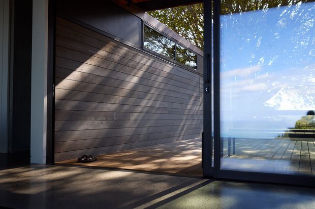 A contemporary indoor-outdoor threshold with a large glazed sliding door opening onto a timber deck. The interior features a dark polished concrete floor and a horizontal timber-clad wall, while the glazing reflects trees and reveals a bright water view beyond. Strong sunlight creates patterned shadows across the floor and wall.