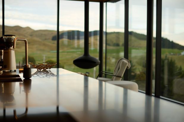 A contemporary open-plan kitchen or dining area with a large glossy light-coloured island or table in the foreground, a suspended black pendant fireplace or feature light, and floor-to-ceiling glazing. Dark slender window frames create expansive views toward rolling green hills and a rural landscape. Minimal furnishings and warm natural light give the space a refined, calm atmosphere.