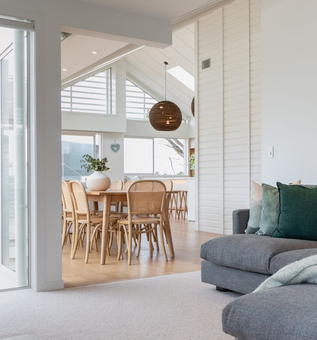 Bright open-plan coastal-style interior viewed from a carpeted living room toward a dining area. The space features a grey sectional sofa with teal cushions, a timber dining table surrounded by woven cane chairs, pale timber flooring, white horizontal wall panelling, large windows, skylights, and a wooden spherical pendant light. The dining area has a high, light-filled ceiling and glazing with outdoor views.