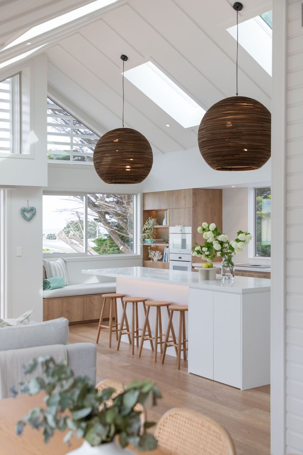 Bright open-plan kitchen and living area with a double-height white pitched ceiling, multiple skylights, expansive windows, and two large woven timber pendant lights. The kitchen features a white island with integrated cabinetry and seating, light timber flooring and cabinetry, a built-in oven, and a window-side banquette. Pale upholstered living furniture and natural floral styling create an airy, warm interior.