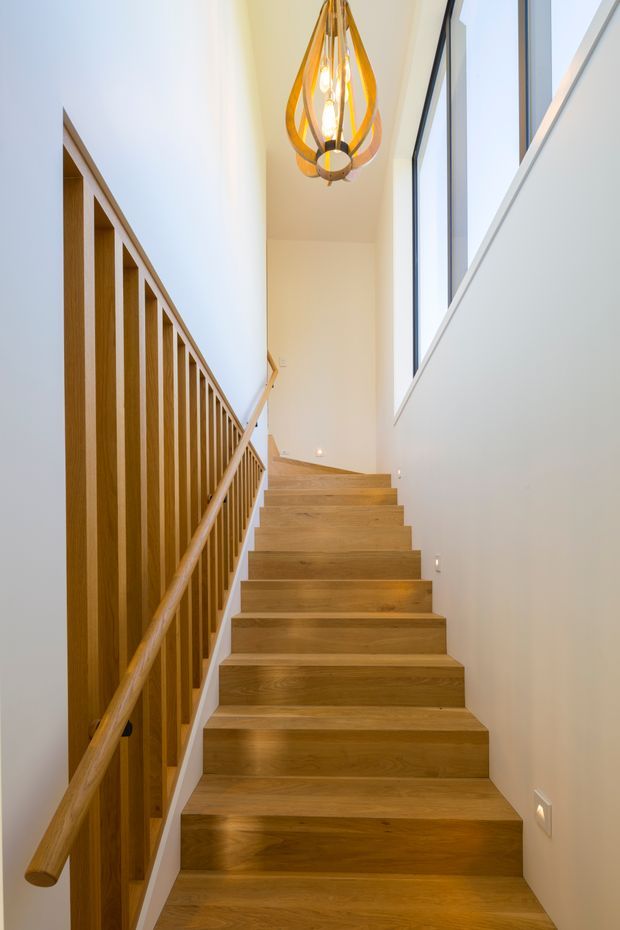 Bright contemporary stairway rising through a narrow double-height passage. The staircase has warm timber treads and matching timber handrail, with a timber vertical-slat balustrade on the left. White walls, tall black-framed windows, recessed step lights, and a sculptural pendant light create a clean, modern interior.