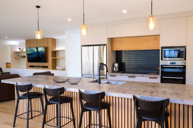 Open-plan contemporary kitchen and living area dominated by a large stone-topped island with seating for five or more. The kitchen features light cabinetry, a dark horizontal-tile splashback, integrated appliances, timber accents, vertical timber screening on the island and feature wall, pendant lighting, and a neutral palette. A living area with television is visible beyond the island.