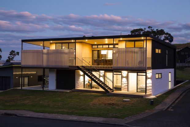 Twilight exterior view of a contemporary multi-storey residence on a sloping suburban site. The house has a box-like form with dark cladding, warm timber-toned illuminated interiors, extensive glazing, upper-level balconies with vertical privacy screens, and an exposed external stair connecting the levels.