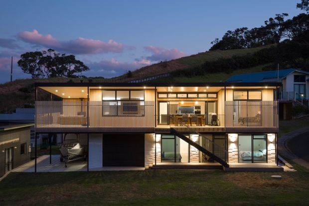 Twilight exterior view of a contemporary, multi-level hillside home. The long rectangular facade features extensive glazing, warm interior lighting, vertical timber-toned screening, dark framing, a covered upper-level terrace or balcony, and an external stair connecting the levels. A garage and parked vehicle are visible below, with a sloped landscaped site and trees behind.