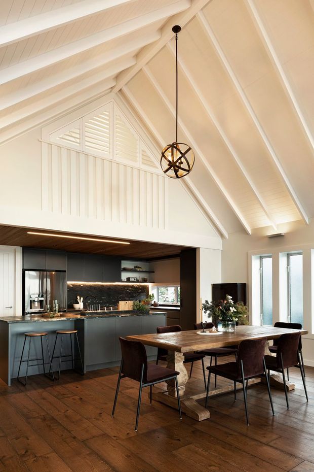 A bright open-plan dining and kitchen interior beneath a dramatic white pitched ceiling with exposed rafters and a decorative gable vent. The room features a large rustic timber dining table with dark upholstered chairs, dark contemporary kitchen cabinetry, a stone-look benchtop, timber ceiling detailing, and dark timber flooring. A spherical pendant light hangs above the dining table, while tall windows provide abundant natural light.