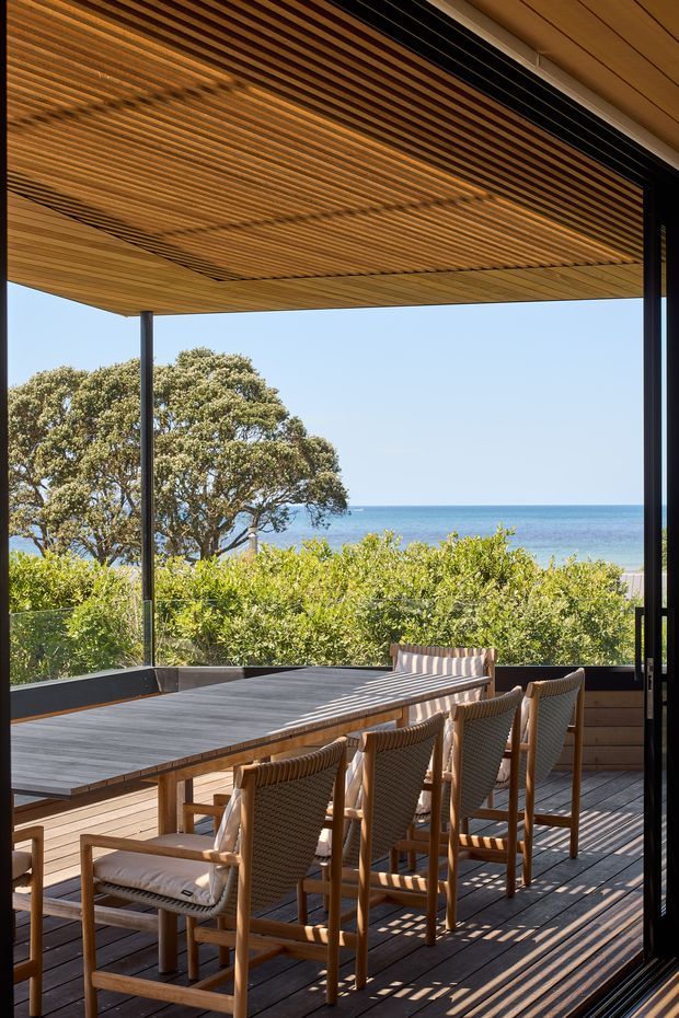 A covered outdoor dining terrace overlooking the ocean. A long dark dining table with woven timber-framed chairs sits on a timber deck, enclosed by slim dark-framed glazing or screens. The ceiling features extensive linear timber battens, creating strong rhythmic shadows and a warm, contemporary coastal atmosphere. Native vegetation and a blue sea view are visible beyond the terrace.