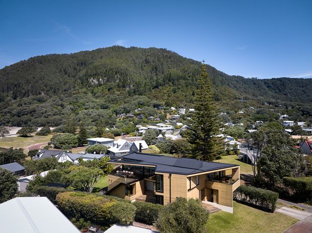 Aerial view of a contemporary single-storey house with warm timber-clad walls, dark low-pitched roofing, extensive glazing, and projecting roof forms. The home sits within a landscaped suburban setting, with a densely forested mountain rising behind the surrounding neighbourhood.