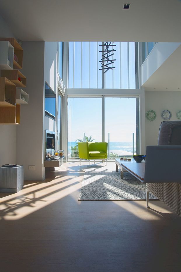 Bright contemporary living room with double-height glazing overlooking the ocean. The room features pale timber flooring, built-in timber shelving and media joinery, a vivid lime-green lounge chair, a grey sofa, a white coffee table, and a sculptural hanging light. Strong sunlight casts long shadows across the floor, creating an airy coastal atmosphere.