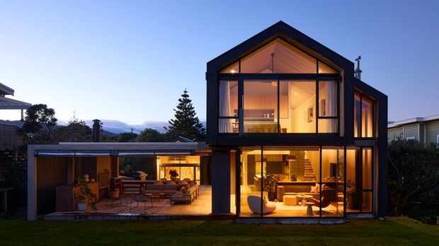 Twilight exterior view of a contemporary multi-level residence with a prominent steep gable form, dark framed glazing, warm timber-lined interiors, and extensive floor-to-ceiling windows. A covered outdoor living area connects to the main house, while surrounding vegetation and distant hills form the background.