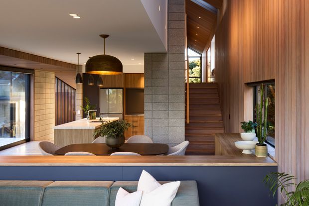 A contemporary open-plan interior combining a living area, dining space, and kitchen, viewed toward a central staircase. The room features extensive warm timber wall panelling and ceiling lining, a grey masonry feature wall, dark blue built-in joinery, a light stone kitchen island, large glazed openings, and sculptural pendant lighting. The staircase rises beside the masonry wall to a brighter upper level.