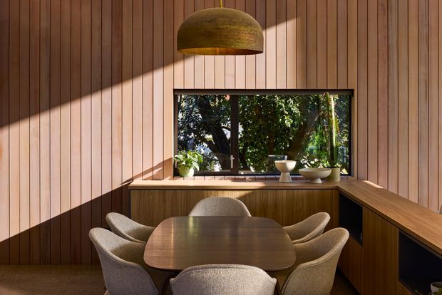 A warm, compact dining area clad in vertical timber wall panelling, with a dark rounded dining table surrounded by upholstered chairs. A large bronze dome pendant hangs above, while a timber built-in sideboard and bench-like joinery frame a black-framed window overlooking greenery. Strong sunlight creates dramatic diagonal shadows across the timber surfaces.