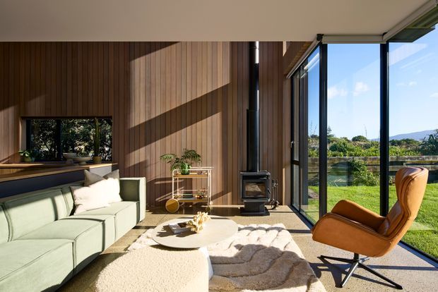 A bright, contemporary living room with full-height glazing opening onto a green garden landscape. The room features warm vertical timber wall panelling, a black freestanding wood-burning stove, a pale green sectional sofa, a sculptural tan leather lounge chair, and layered neutral rugs around a low oval coffee table. Strong sunlight creates dramatic shadows across the timber feature wall and floor.