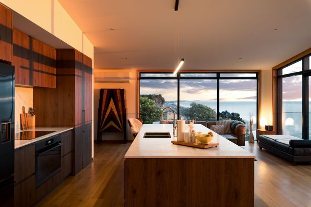 A warm, open-plan kitchen and living area with a large central island, dark timber cabinetry, integrated appliances, and a pale stone benchtop. Full-height black-framed glazing opens to expansive coastal or waterfront views, while a dark sofa and lounge furnishings sit beyond the kitchen. Linear pendant lighting, timber flooring, and dramatic evening light create a refined contemporary atmosphere.