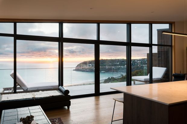 Contemporary open-plan interior with full-height black-framed glazing overlooking a coastal bay and headland at sunset. A minimalist living area with a low dark sofa, lounge chairs, timber flooring and a large kitchen island occupies the foreground. The space has a warm, refined atmosphere and strong indoor-outdoor visual connection.