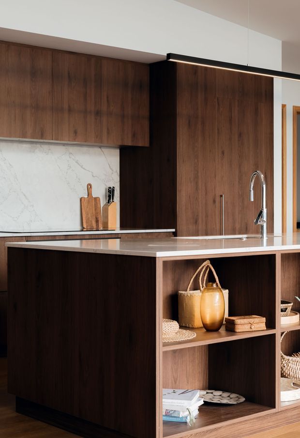 A refined contemporary kitchen dominated by dark walnut-toned cabinetry and a large central island. The island has a pale stone benchtop with an integrated open shelving display containing baskets, books, pottery and serving pieces. Matching timber wall cabinetry, a white veined stone splashback, chrome gooseneck tap, timber chopping boards and a slim black linear pendant light complete the warm, tailored fit-out.