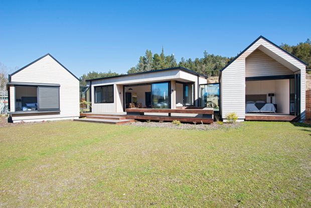 Exterior view of a contemporary low-rise residential compound composed of three separate gabled and flat-roofed volumes. The central pavilion has dark-framed glazing, a timber deck and a sheltered outdoor sitting area, while the side buildings feature white horizontal cladding and large openings. The right-hand volume visibly contains a bedroom. The buildings sit in a landscaped grassy setting with trees behind, under a clear blue sky.