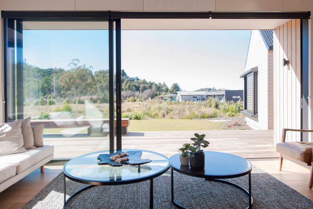 Bright contemporary living room opening onto a spacious timber deck through wide floor-to-ceiling glazed sliding doors. The room features a light upholstered sofa, two round black-framed coffee tables with glass tops, a textured dark rug, and a timber-clad exterior wall visible beside the deck. The outlook includes landscaped surroundings and neighbouring buildings.