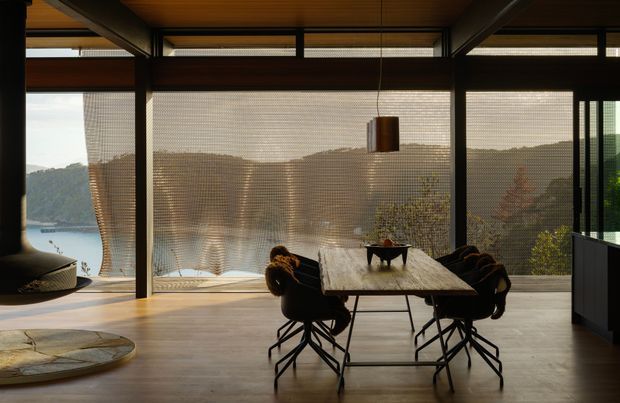 A contemporary dining area with a large timber dining table and dark upholstered chairs, set within a glazed living space overlooking a wooded waterfront landscape. The room features exposed dark structural beams, a warm timber ceiling, broad floor-to-ceiling glazing with woven roller blinds, polished concrete flooring, and a sculptural pendant light. A partial lounge area and low built-in cabinetry are visible at the edges.