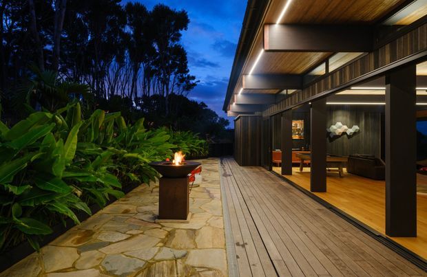 Twilight exterior view of a contemporary home opening onto a wide timber deck. The dark timber-clad facade has deep roof overhangs, exposed soffit lining, recessed linear lighting, and large glazed sliding doors revealing a warm interior living area. A natural irregular-stone-paved terrace runs alongside lush tropical landscaping and a small outdoor fire feature.