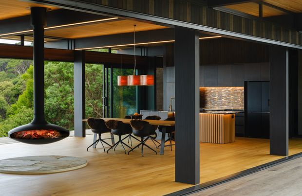 A refined open-plan contemporary interior with a dining area and kitchen opening onto a lush wooded landscape. The space features warm timber flooring and ceiling lining, dark structural steel posts and framing, extensive floor-to-ceiling glazing, a central suspended black fireplace, a long timber dining table with dark upholstered chairs, and a dark kitchen with a textured metallic-toned splashback and ribbed island cabinetry. Linear integrated lighting and a vivid pendant add visual interest.