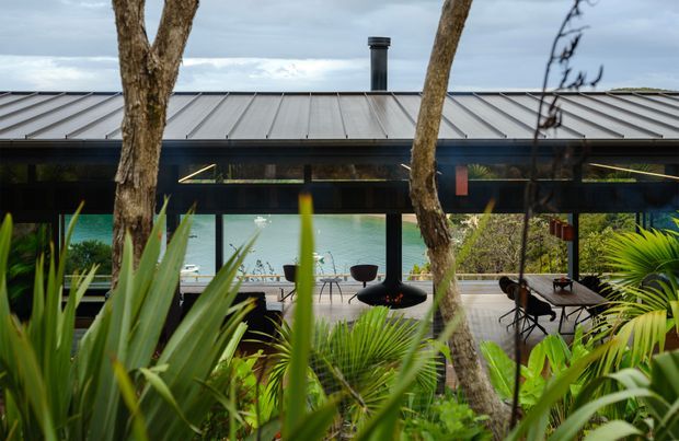 A low, modern pavilion-style house viewed through lush tropical landscaping. The building has a broad grey metal roof, dark expansive glazing and a central chimney, with a sheltered outdoor terrace overlooking turquoise water. Outdoor chairs and tables are visible beneath the deep roof overhang, while mature trees and palms frame the foreground.