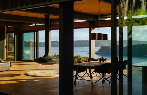 A refined open-plan living and dining interior with extensive floor-to-ceiling glazing overlooking a calm waterfront landscape. The room features warm timber flooring and ceiling lining, dark structural columns and window frames, a suspended sculptural fireplace, a dining table with contemporary chairs, and strong indoor-outdoor visual connection. The image has a warm, dramatic, high-contrast atmosphere.