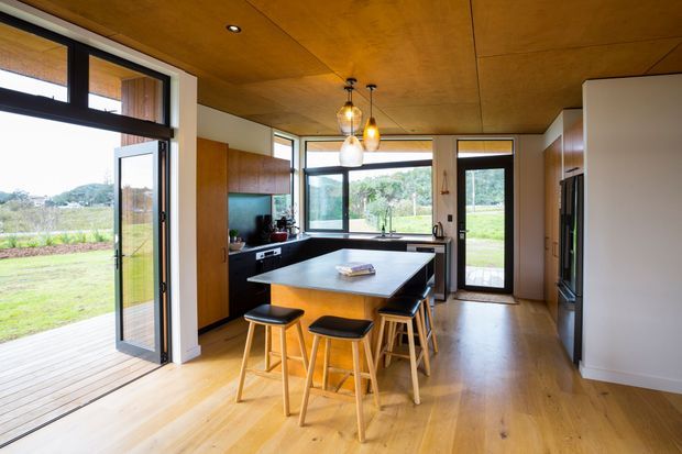 A warm, open-plan contemporary kitchen with a central island and seating for four, surrounded by extensive black-framed glazing and doors opening onto a timber deck and green landscape. The room features a timber-lined ceiling, light timber flooring, dark kitchen cabinetry, white walls, integrated appliances, and pendant lighting over the island.