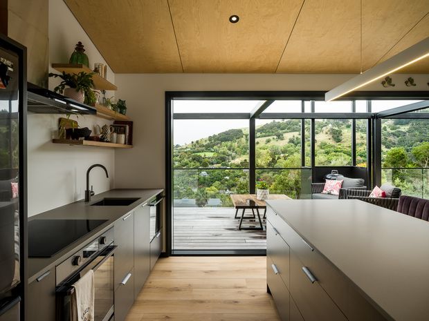 Contemporary kitchen with a large central island, black benchtops, light cabinetry, timber flooring and a warm plywood ceiling. Full-height black-framed glazing opens onto a timber deck and overlooks a green hilly landscape. Open shelving, integrated appliances and pendant lighting complete the space.