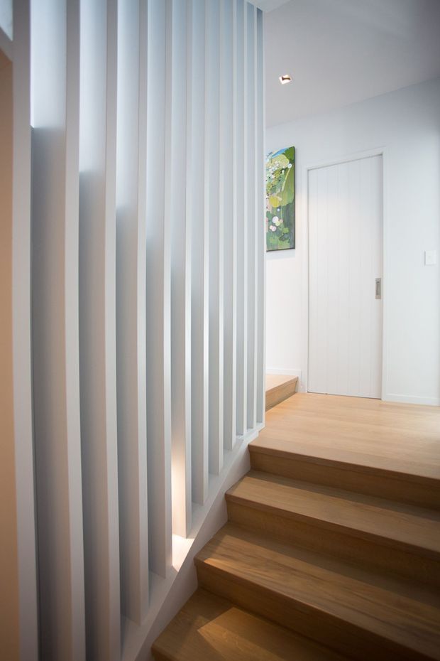 A bright, minimalist interior stairway with warm timber steps and landing, enclosed by a dramatic vertical white slatted screen/partition. White walls and a white paneled door create a clean modern palette, with recessed lighting and a small colourful artwork adding visual interest.