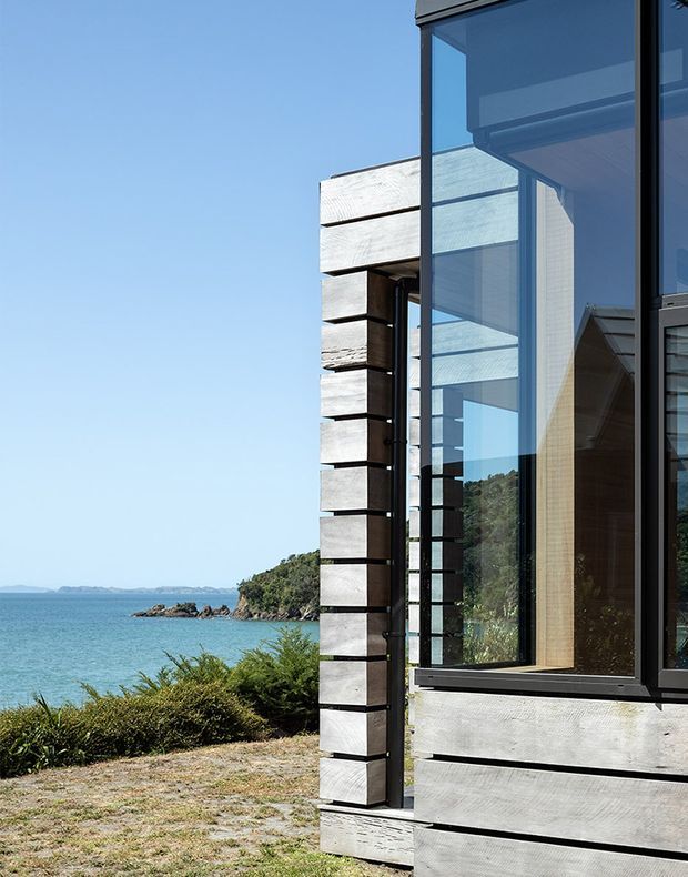 Close exterior view of a contemporary coastal house beside a blue waterfront. The facade combines broad horizontal weathered timber cladding with dark framed floor-to-ceiling glazing and projecting timber elements. A rocky, vegetated shoreline and clear sky are visible beyond the building.