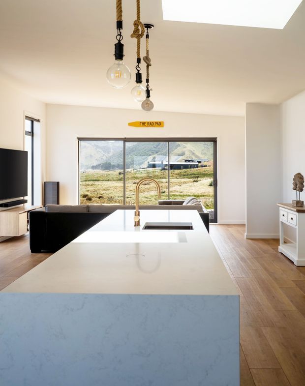 Bright contemporary open-plan kitchen and living area viewed from behind a large island. The kitchen features a pale stone benchtop with an integrated sink and brass-toned tap, light blue island cabinetry, timber flooring, and pendant lights suspended from ropes. A black sofa, television, console, and sideboard occupy the living area, while large sliding windows provide a view toward a rural mountain landscape. A large skylight adds strong natural light.