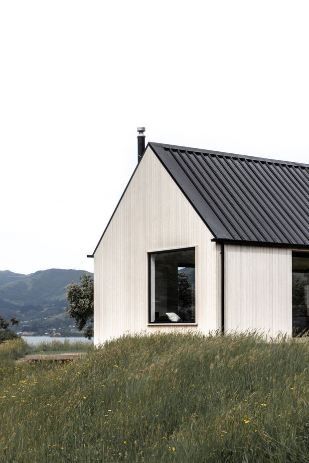 Exterior view of a compact gabled house set in a meadow, with pale vertical cladding, a dark corrugated pitched roof, black-framed windows, and a chimney. A mountainous landscape and body of water are visible in the background.