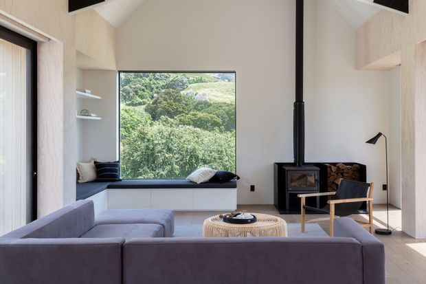 A bright contemporary living room with a high sloping ceiling, pale timber wall panelling, built-in window seating and shelving, and a large picture window overlooking dense greenery. A grey sectional sofa faces a woven round coffee table and a black wood-burning stove, with a timber-framed lounge chair and floor lamp completing the room.