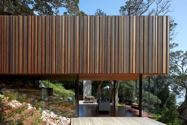 Exterior view of a contemporary elevated home set among trees, with a prominent upper facade clad in closely spaced vertical timber slats. The lower level is largely enclosed in floor-to-ceiling glazing, revealing a living area and dining setting, while a timber deck projects in front. Natural rocky landscaping and mature trees surround the building.
