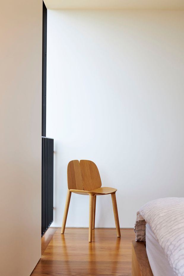 A minimalist bedroom corner with a warm timber floor, a simple sculptural wooden chair, and the edge of a bed with pale bedding. A tall black vertical radiator or grille contrasts with the white walls and natural wood tones.
