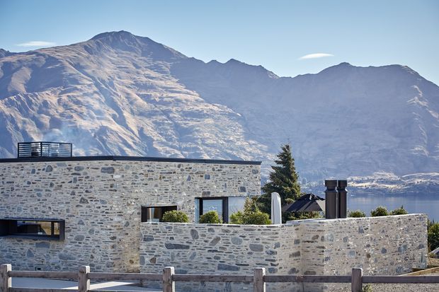 Exterior view of a contemporary, low-profile stone-clad house set against dramatic mountains and a lake. The building has flat rooflines, extensive grey natural-stone walling, dark-framed windows, rooftop terrace railings, and a sheltered outdoor area. A timber fence is visible in the foreground, with distant mountains and water forming the background.
