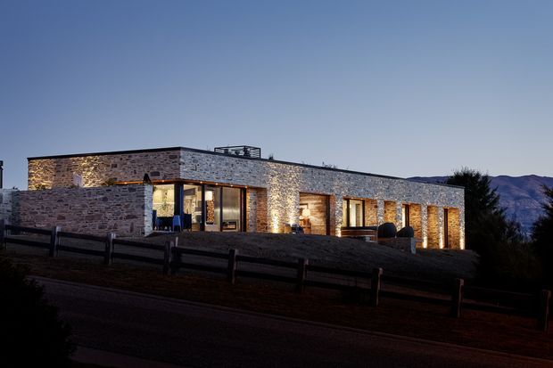 A low, single-storey modern house photographed at dusk, with long horizontal stone-clad walls, a flat roof, and warm light glowing from recessed window and door openings. The building sits in a mountainous setting behind a dark timber fence, with exposed masonry and carefully illuminated exterior surfaces creating a refined, dramatic appearance.
