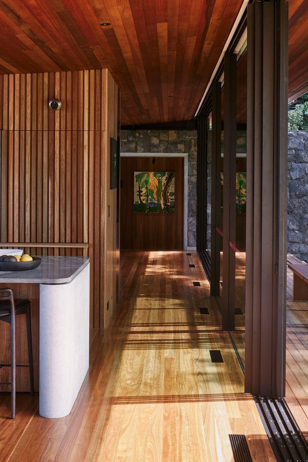 A warmly lit modern interior corridor beside a compact kitchen or bar area. Rich timber boards cover the ceiling, floor, and slatted wall, while full-height dark-framed glazing runs along the right side, opening toward a stone exterior wall and deck. A polished dark stone benchtop, pale curved cabinetry, timber stool, built-in wall detailing, and framed artwork are visible. Strong linear shadows from the glazing create a graphic pattern across the timber floor.
